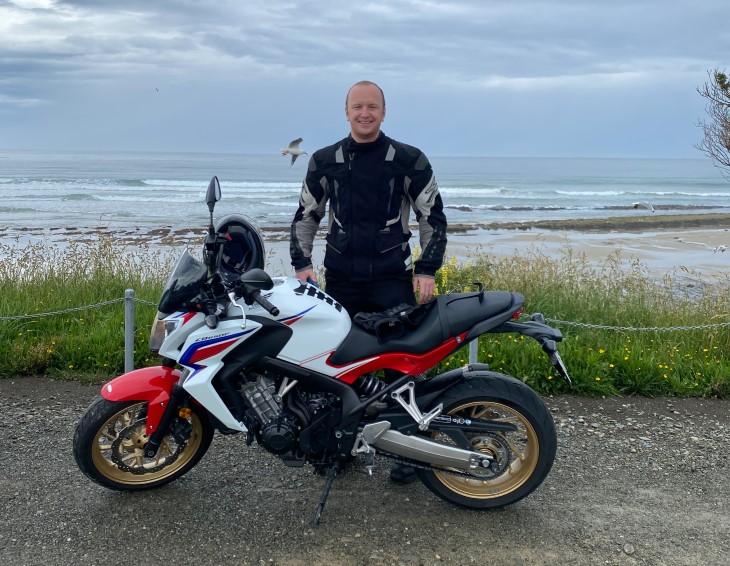 Nick Grant standing with his motorbike on a road in front of the beach. 
