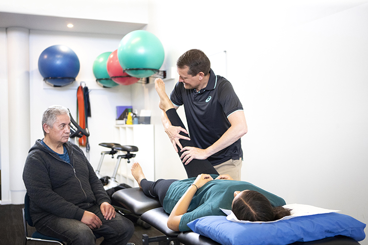 physio cultural safety v2 A young woman receiving physio treatment on her leg while an older man looks on.
