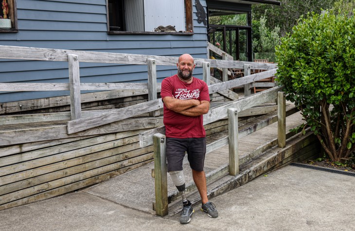 Derek leans against his deck, arms crossed, in a red t-shirt smiling for the camera