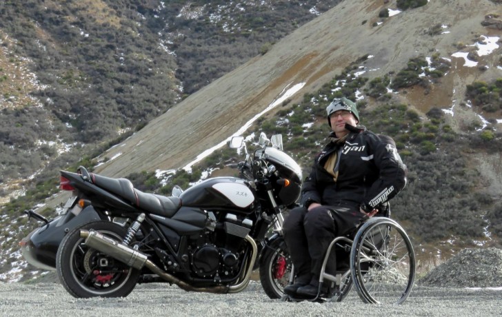 Brett Ladbrook sitting in his wheelchair in front of his motorbike with a mountain behind him.