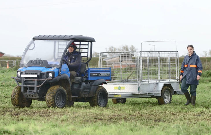 A male farmer driving a quad vehicle towing the new calf trailer while a female colleague walks alon