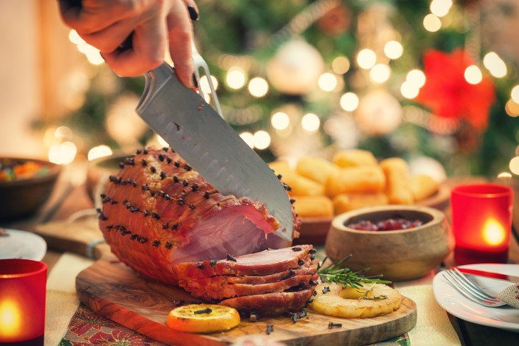 A close-up shot of a ham being carved into slices on a table with a Christmas tree in the background. 