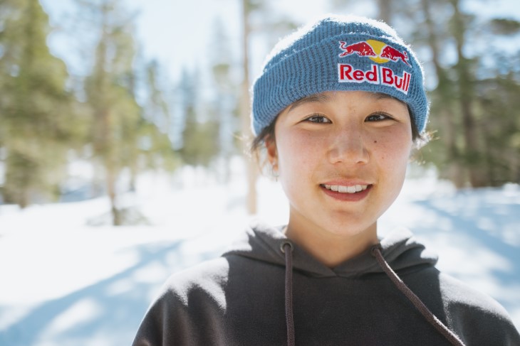 A close-up photo of Cool Wakushima's face, she's wearing a beanie with snow and trees behind her in the background.