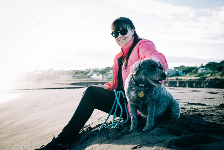 Diane Alder sitting on the beach with her dog and smiling at the camera. 