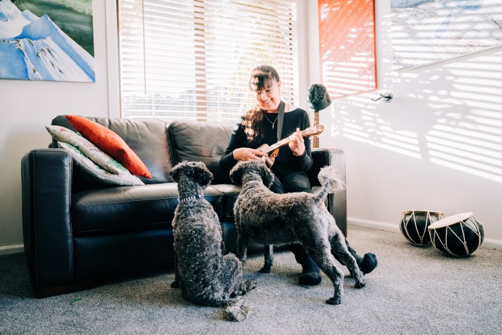 Diane Alder playing the ukelele on her couch in front of her two dogs.