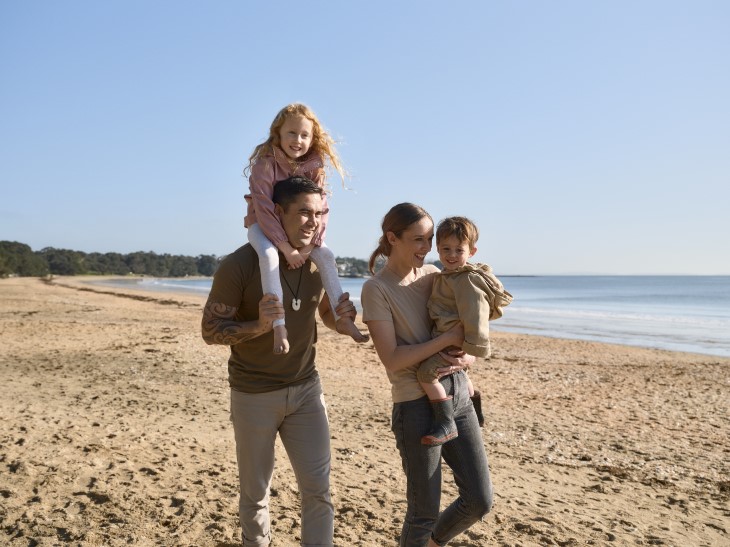 A young couple walking on the beach carrying their two children.