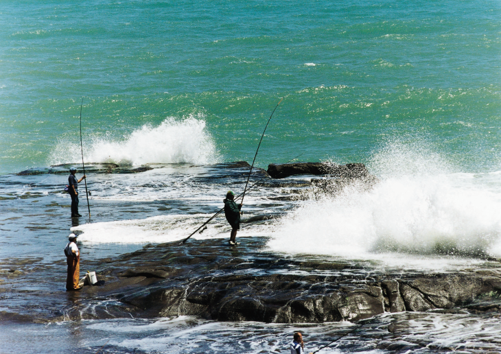 Several people fishing on rocks next to the ocean. 