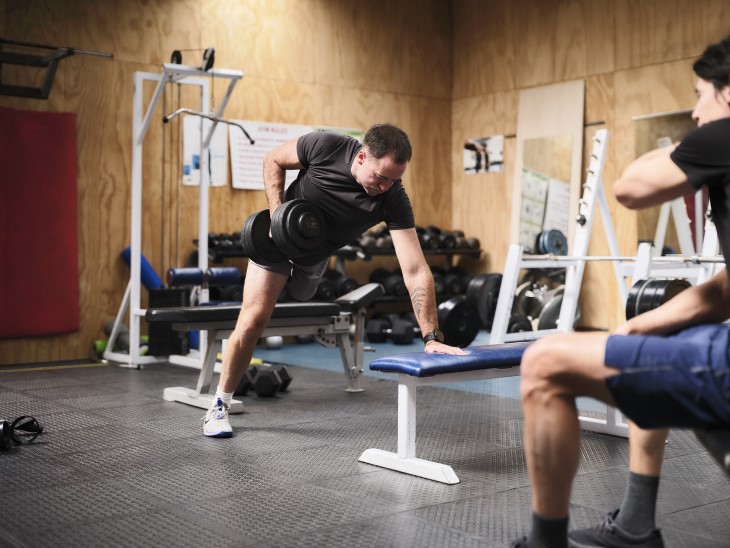 A man lifting a free weight in the gym while another man looks on.