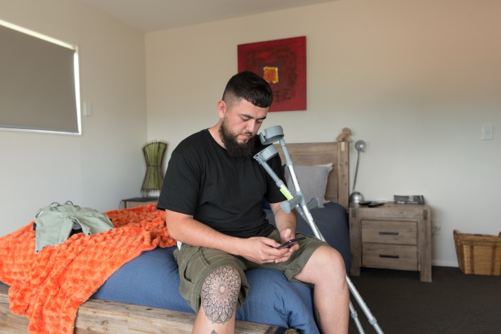 An injured man sitting in his bedroom and looking at his phone, his crutches are leaned up against the bed.