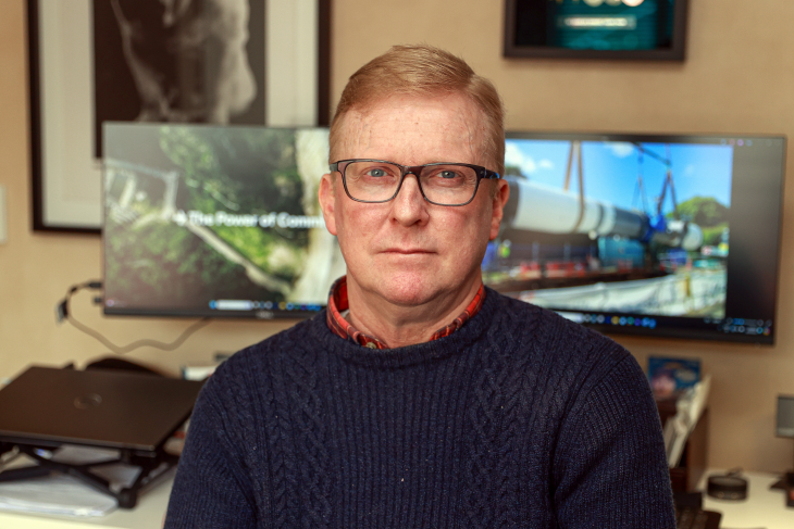 A close-up photo of Jason Ross at home, with his work computers in the background.