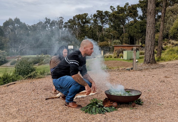 Aboriginal man Jeffrey Gray kneeling down next a plume of smoke coming from plants being heated outs