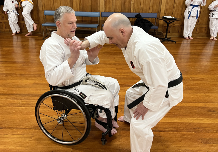 John Marrable doing karate training in his wheelchair with another man, who is standing up.