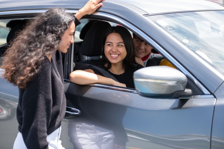 A young woman sitting in a car and talking to her friend, who is standing next to the car.