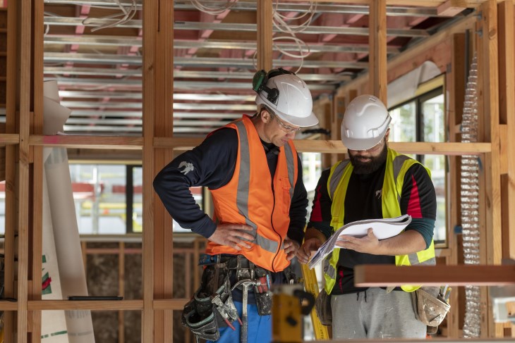 Two builders standing at a building site and looking at a document one of them is holding.