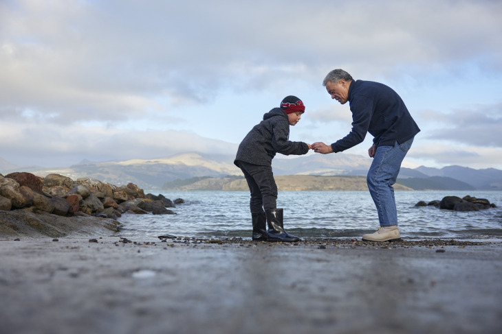 A middle-aged man and a young boy bending down to look at a stone on the beach.