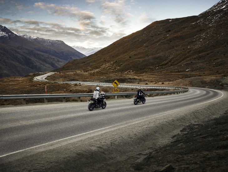 Two motorcyclists riding along a winding road with mountains in the background.