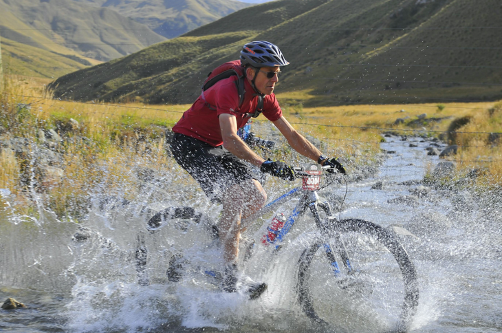 A male mountain biker riding through the water of a shallow river.