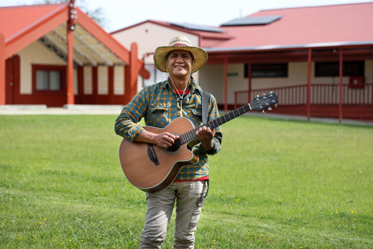 Pita Pene holding his guitar and smiling, with a marae in the background.