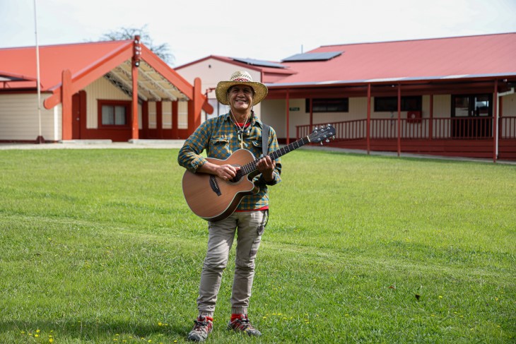 Pita Pene holding his guitar and smiling, with a marae in the background.