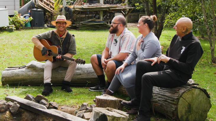 Pita Pene sitting on a log outside with his ACC recovery team and playing his guitar for them. 