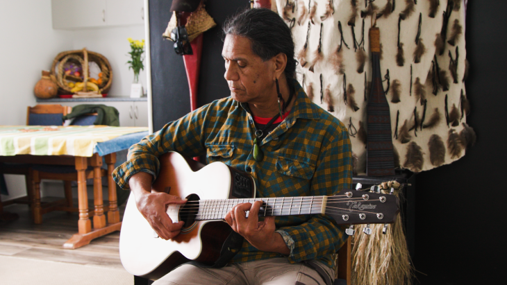 Pita Pene sitting down and playing the guitar in his home.