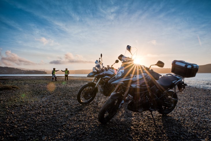 Two motorbikes parked next to the water with their riders in the background. 