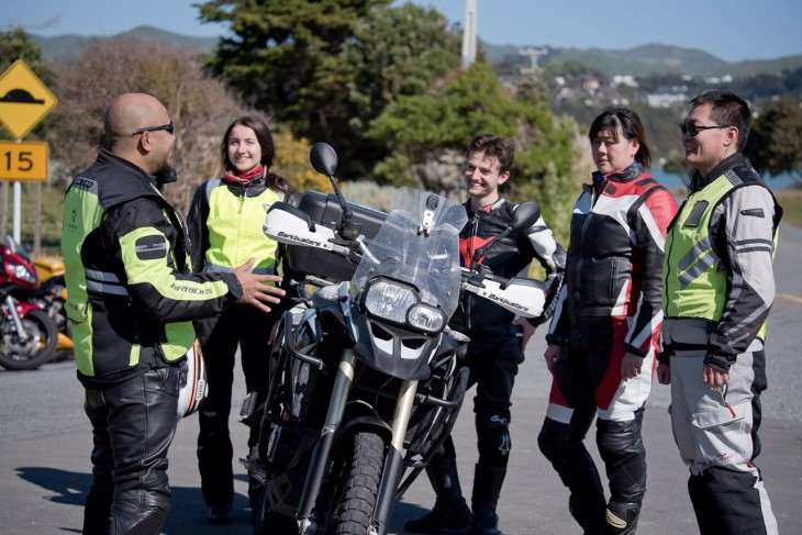 A group of riders standing around a motorbike talking to each other. 