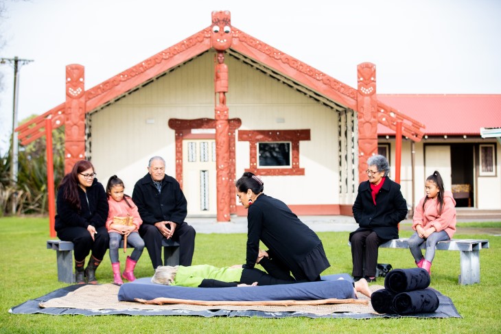 A Rongoā Māori practitioner using her healing methods on a woman while whānau watch on. 