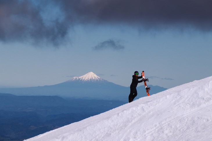 A snowboarder stands on a slope with another mountain behind him in the distance. 