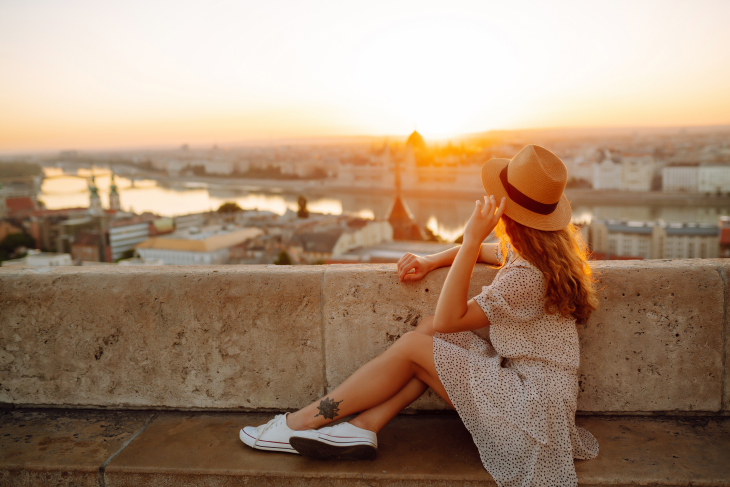 A woman sitting on a rooftop overlooking a sunset at a city in Europe. 