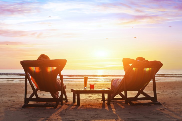 A couple relaxing on deck chairs on a beach watching the sunset.