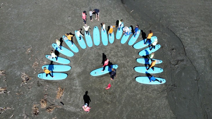 An aerial shot showing surfboards arranged in a semi-circle on the beach with people standing on the