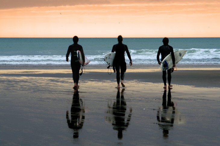 Three surfers shown from behind walking out to the water at a beach. 