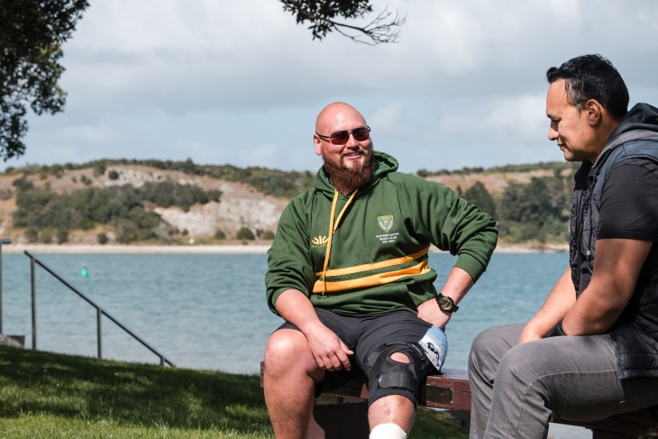 Taane Karena sitting on a bench on the coast with his ACC recovery partner Lance Kilgour.