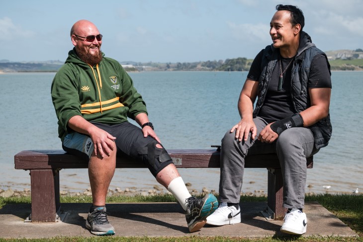 Taane Karena sitting on a bench on the coast with his ACC recovery partner Lance Kilgour, both are laughing.