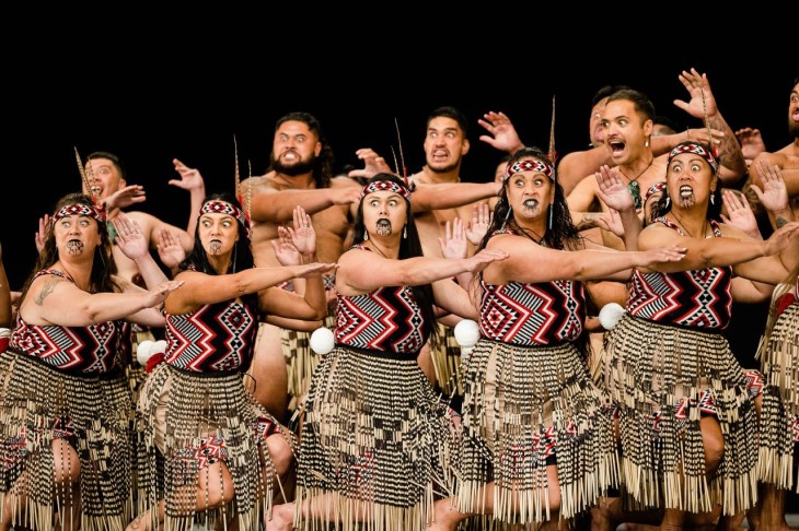 A group of men and woman wearing traditional Māori clothing and performing kapa haka.