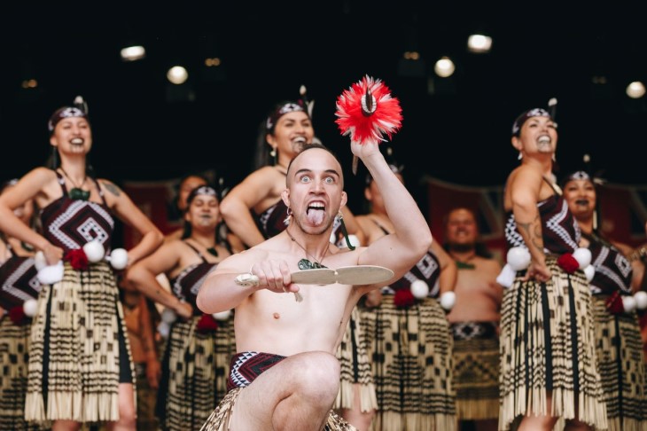 A man kneeling down with other members of his group behind him as part of their kapa haka performance, they're all wearing traditional Māori clothing.