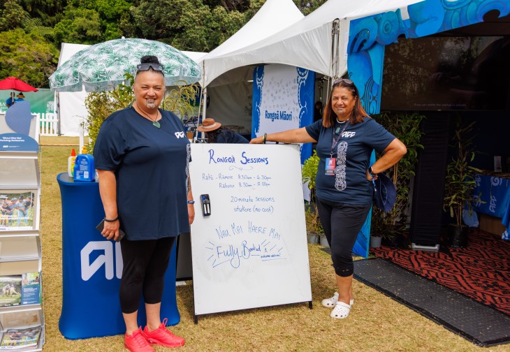 Two female ACC staff members standing next to a whiteboard advertising rongoā sessions outside the ACC tent.