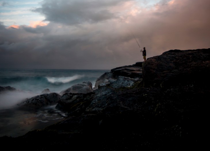 A lone fisherman standing on a rock casting his line out into the ocean. 